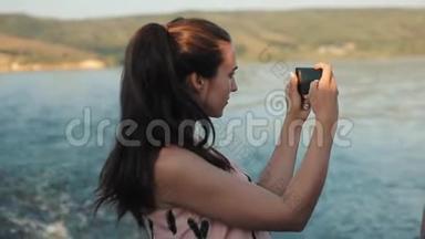 一个梳着马尾辫的漂亮女孩的侧半身景，用她的手机拍下了河和山的照片